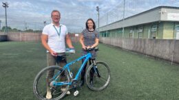 A man and a woman stand behind a blue frame bike on a five-a-side football pitch with a building in the background