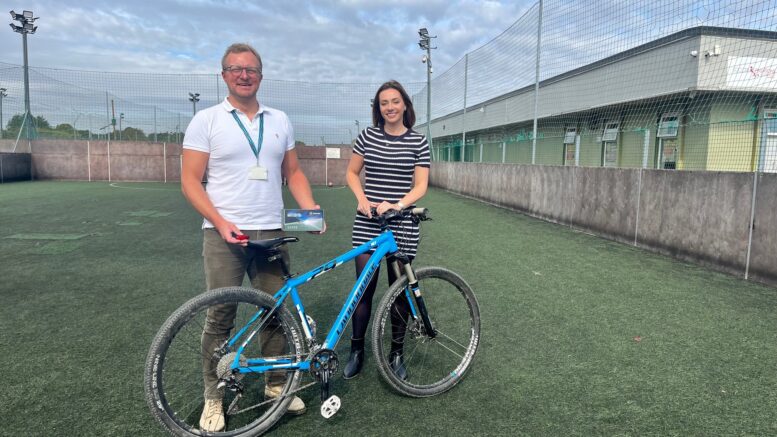 A man and a woman stand behind a blue frame bike on a five-a-side football pitch with a building in the background