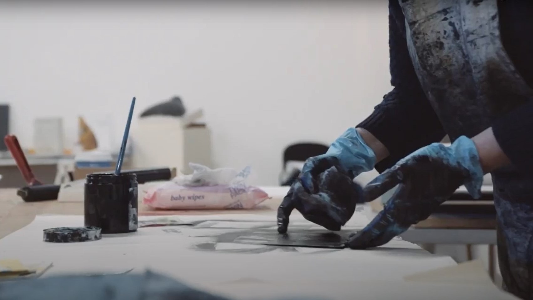 A pair of hands in gloves pick up some printed paper with a pot of paint and other artists tools in background