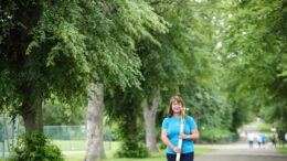 A women in a light blue top holds the gold baton of hope. She stands in a park, with mature trees in the background