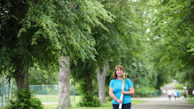 A women in a light blue top holds the gold baton of hope. She stands in a park, with mature trees in the background