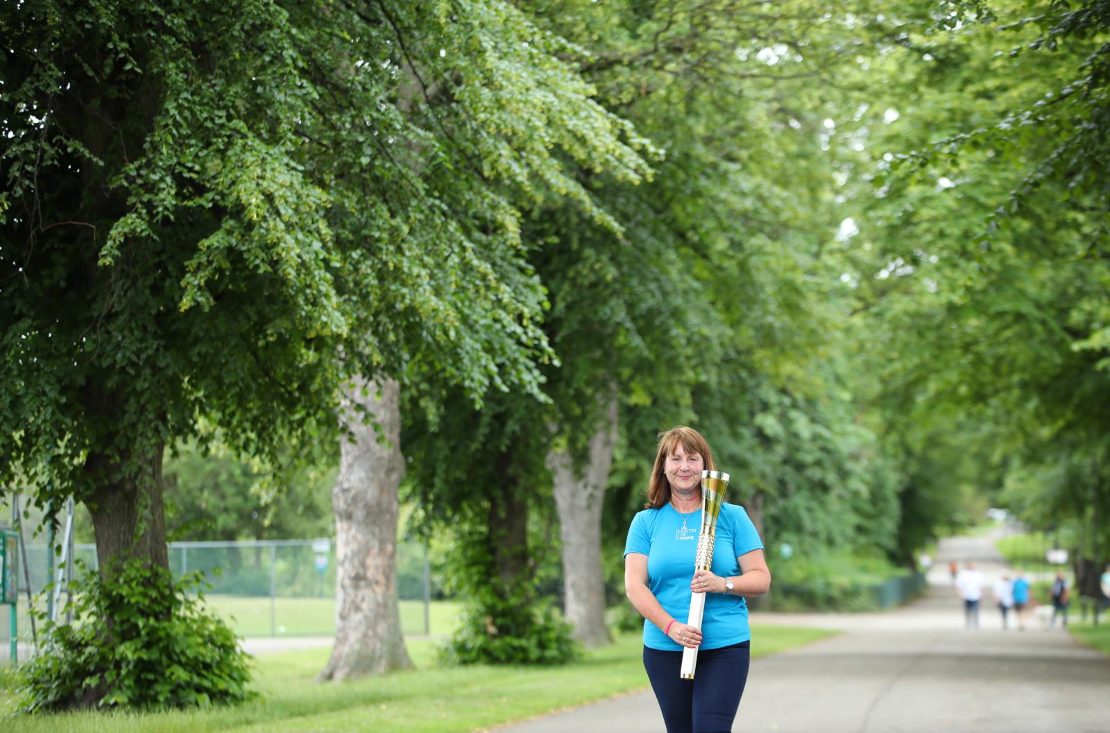 A women in a light blue top holds the gold baton of hope. She stands in a park, with mature trees in the background