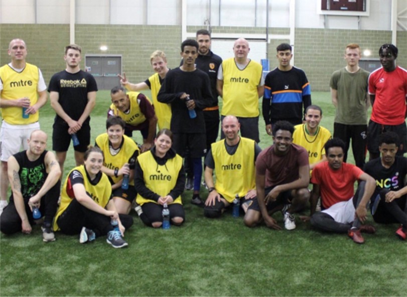 A group of people of various ages on an indoor football pitch pose together - some in yellow bibs.