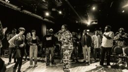 A black and white image of a man in camo clothes with a microphone performing with people behind him watching