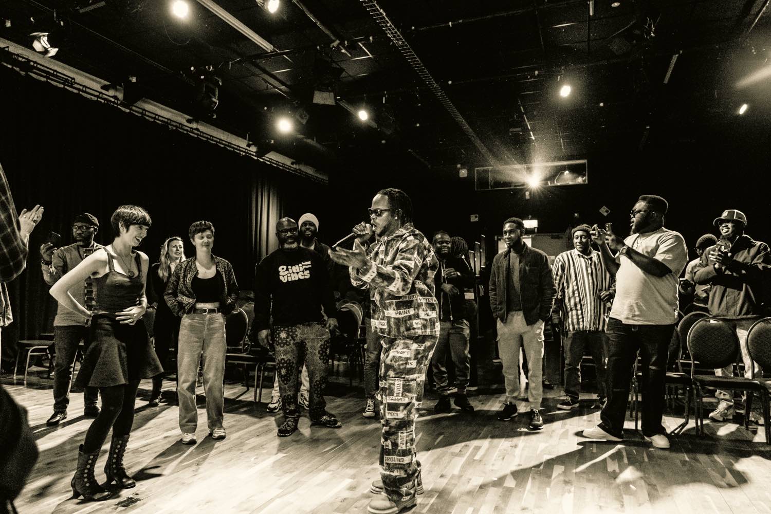A black and white image of a man in camo clothes with a microphone performing with people behind him watching