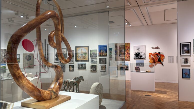 A large bright gallery room with artwork on the walls in the background and a sculpture in a glass case in the foreground.