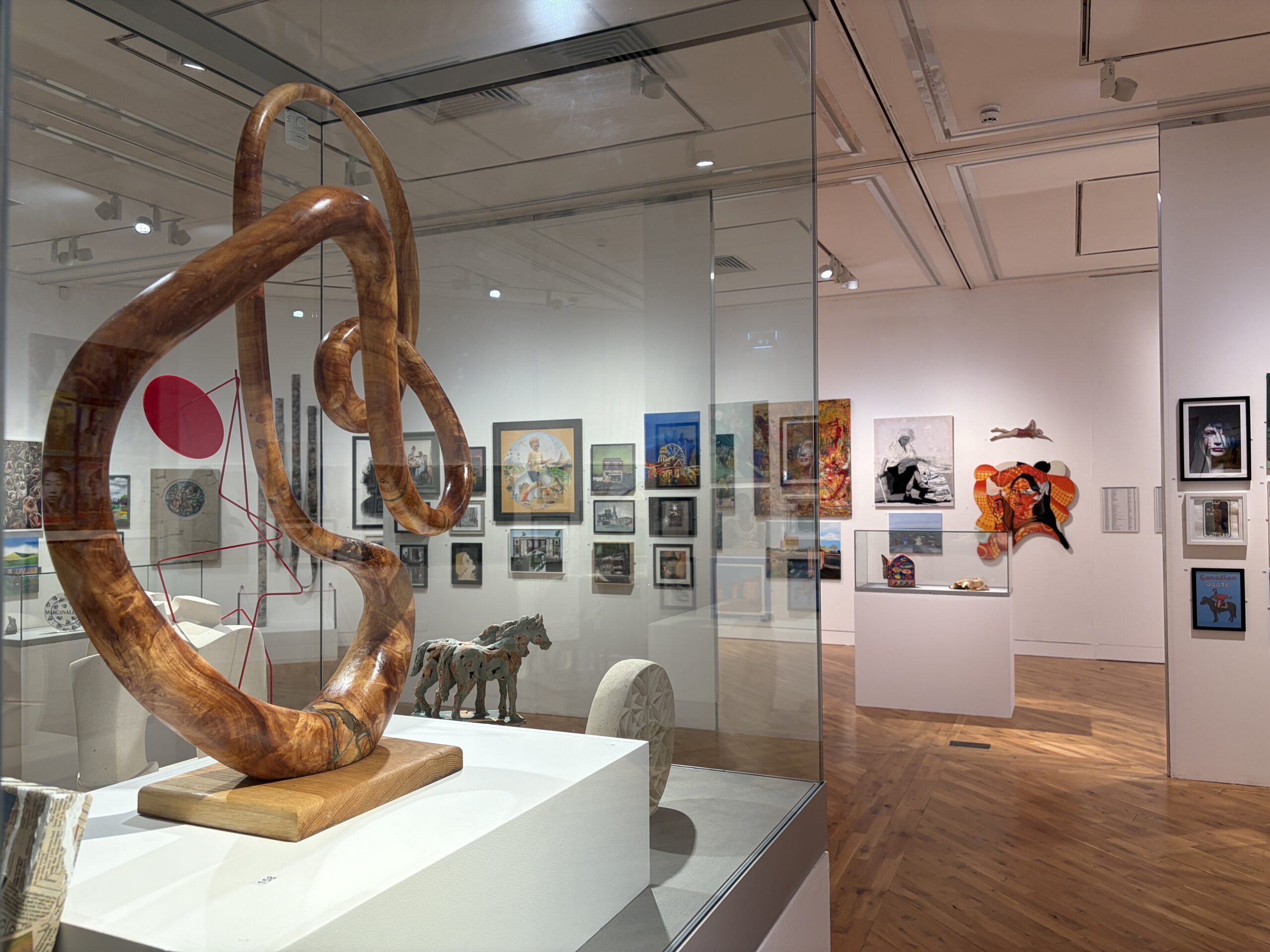A large bright gallery room with artwork on the walls in the background and a sculpture in a glass case in the foreground.