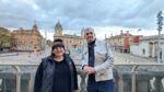 A woman and a man stand on a balcony with a city centre square and monument in the background below
