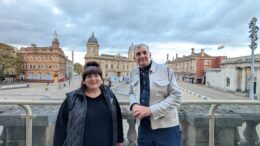 A woman and a man stand on a balcony with a city centre square and monument in the background below