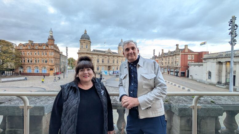 A woman and a man stand on a balcony with a city centre square and monument in the background below