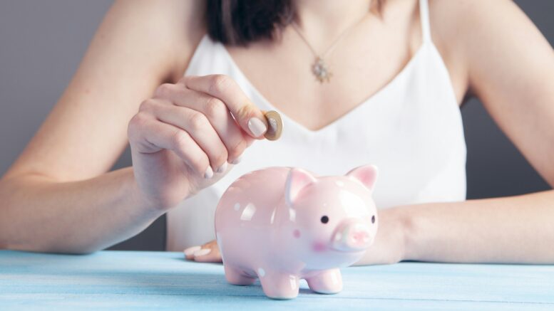 an image of a pink piggybank. A person in the background is placing a £1 coin inside it. We see their hand and torso, but not their face