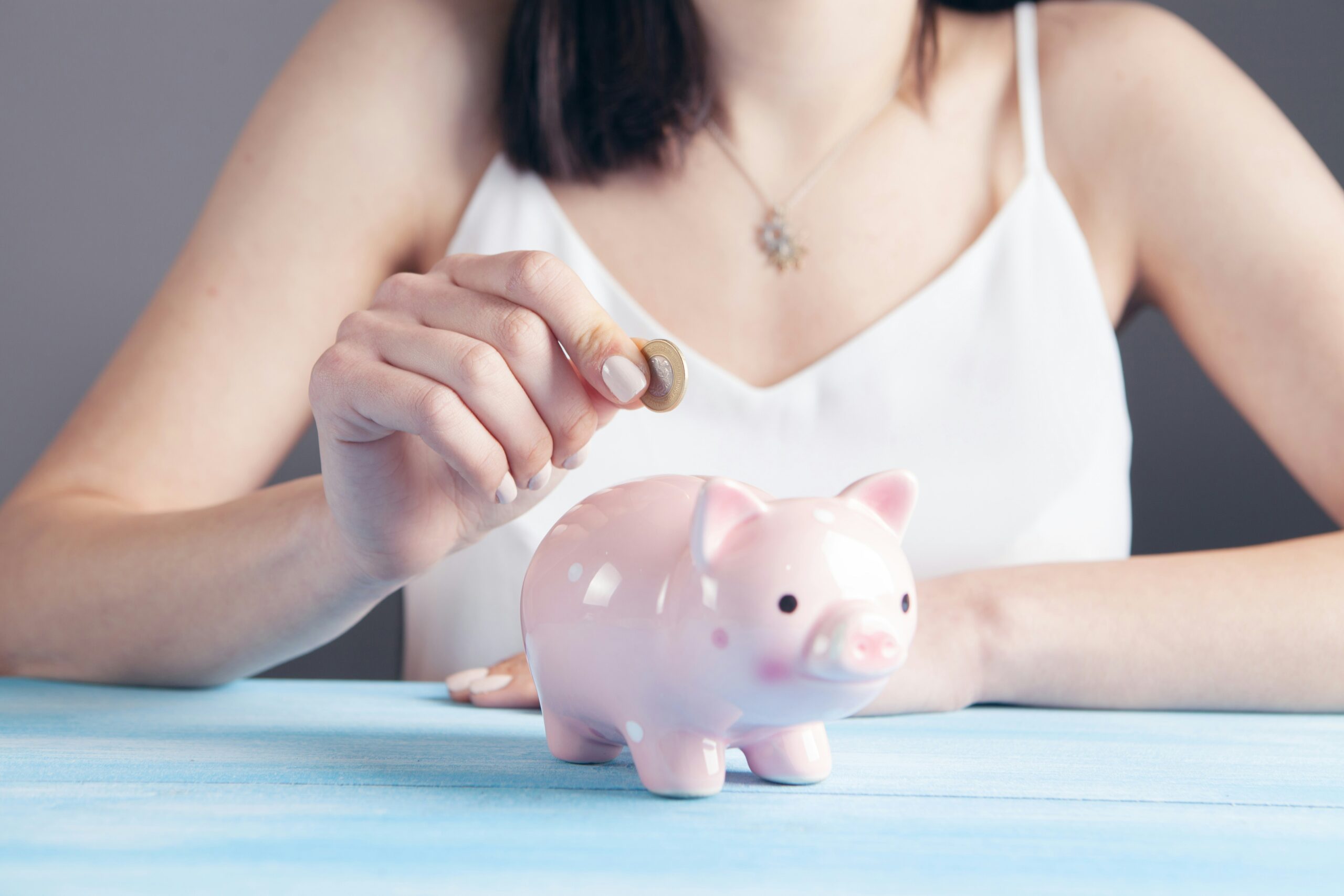 an image of a pink piggybank. A person in the background is placing a £1 coin inside it. We see their hand and torso, but not their face
