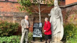 A tall stone statue of Minerva stands in the corner of a garden next to a new black plaque with information and black and white images. a man and woman stand either side of the plaque