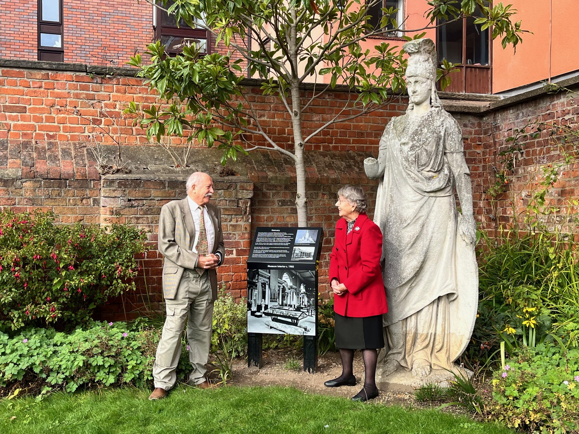 A tall stone statue of Minerva stands in the corner of a garden next to a new black plaque with information and black and white images. a man and woman stand either side of the plaque