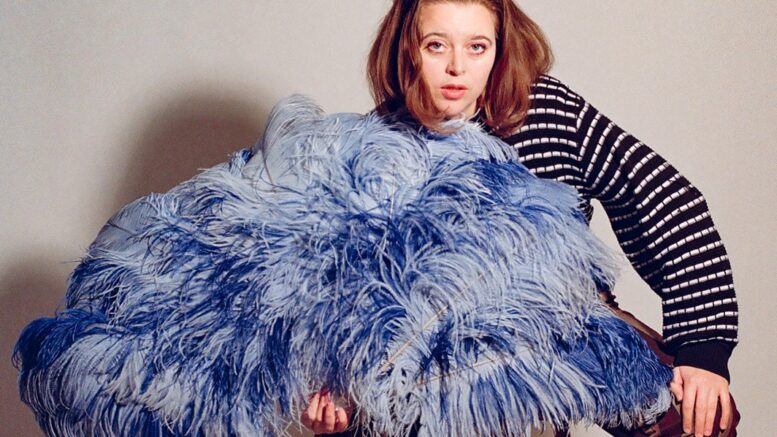 A woman sits on a stool holder a large blue feather fan