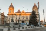 A large historic building with the sunset reflecting on it and a large christmas tree in the foreground.