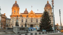 A large historic building with the sunset reflecting on it and a large christmas tree in the foreground.