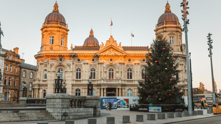 A large historic building with the sunset reflecting on it and a large christmas tree in the foreground.