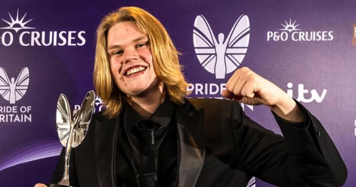 A man with long blonde hair and a black tie suit stands holding a silver trophy in front of a board with Pride of Britain written on it