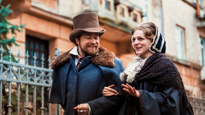A man and a woman in Victorian costume walk arm in arm down a street