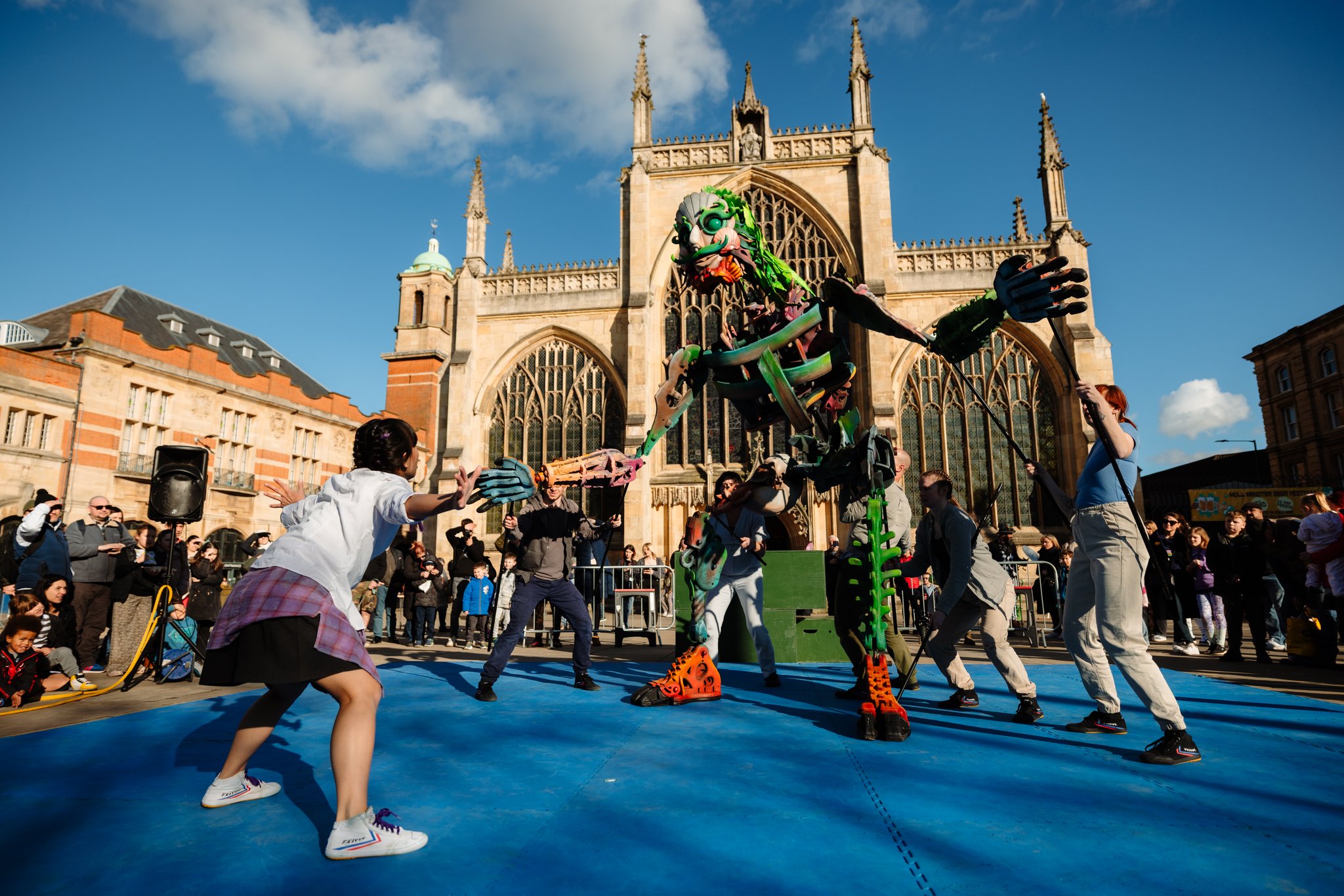 A large, colourful puppet operated by several performers interacts with a child on a blue mat in front of Hull Minster.