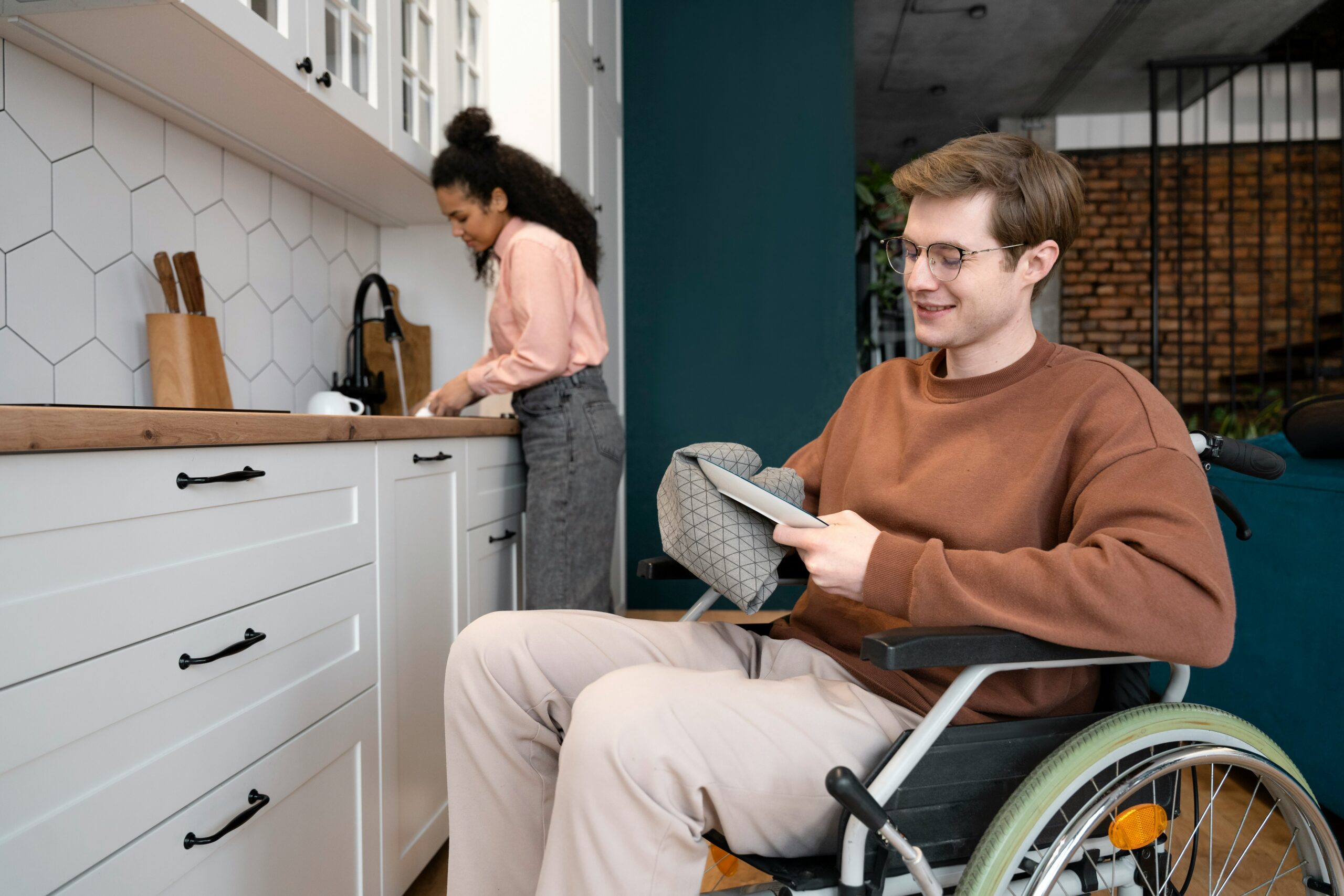 Woman stood by a sink and a young man in a wheelchair in the foreground.