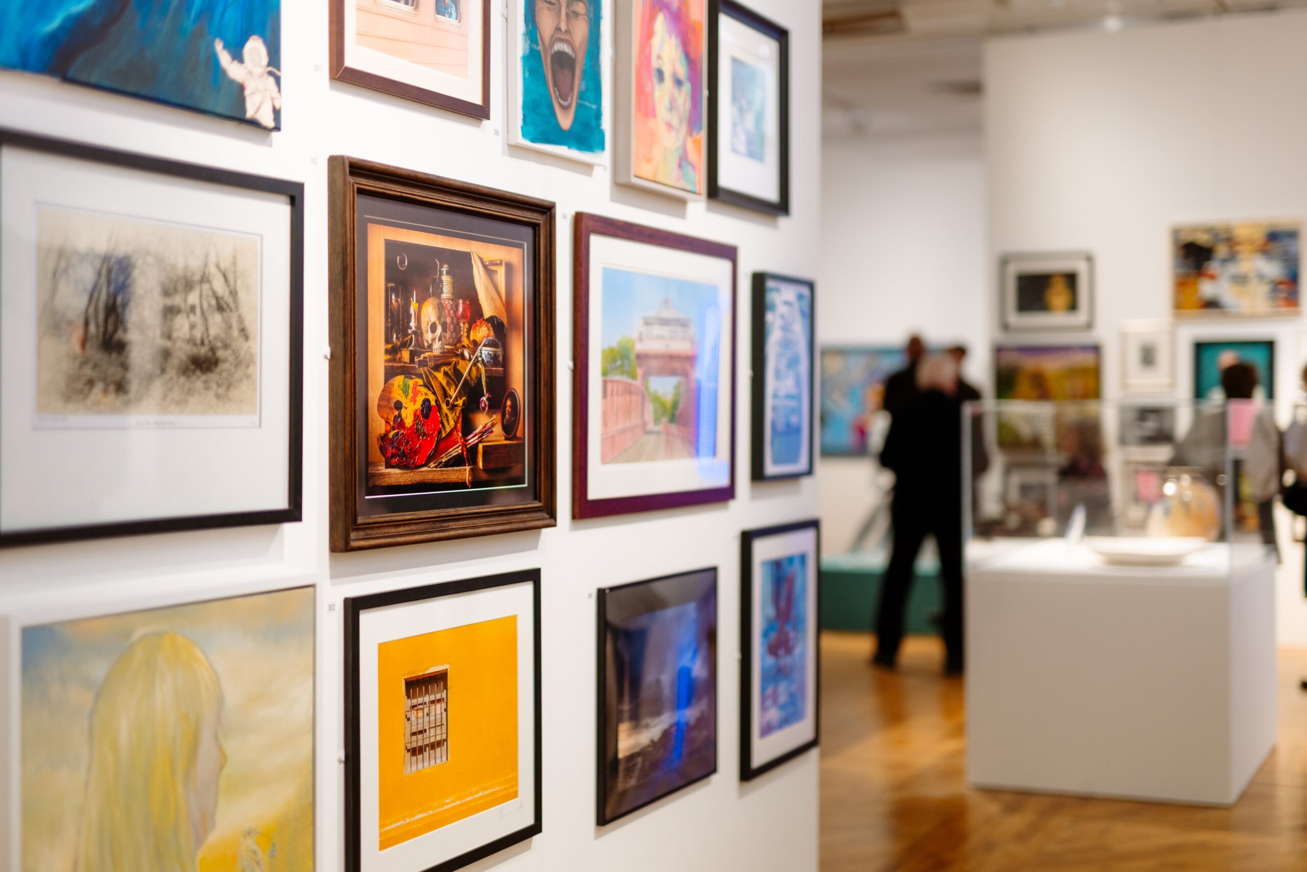 A image of a white gallery wall of paintings in various frames in the forground. Blurred people and further artwork in the background