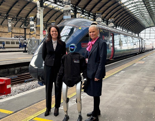In a train station with Hull Trains train in background, and two women stood in front either side of a humanoid robot