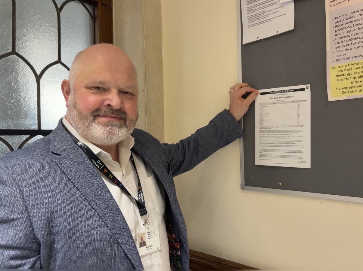 A man wearing a blue suit with a grey beard pins a piece of paper to a noticeboard