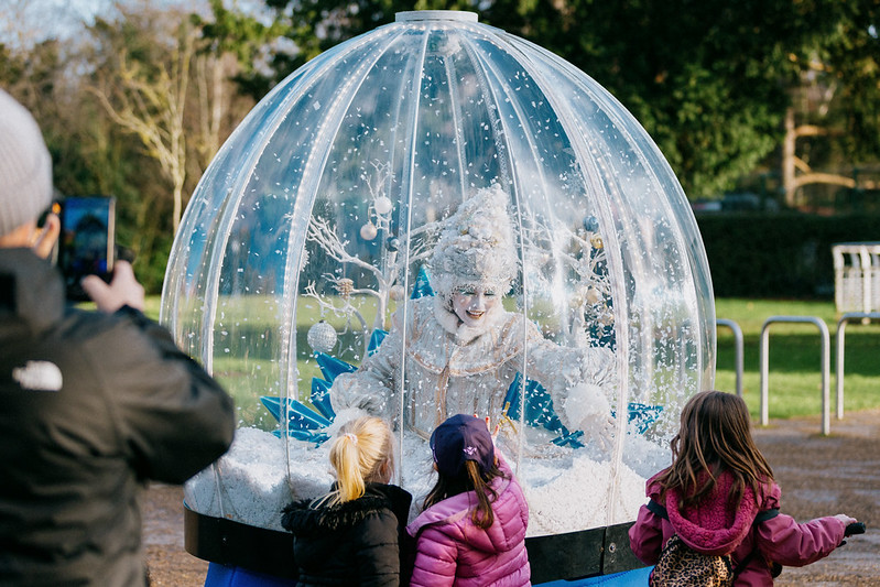 A woman dressed as an ice queen sits within a clear snowglobe with children looking through at her,