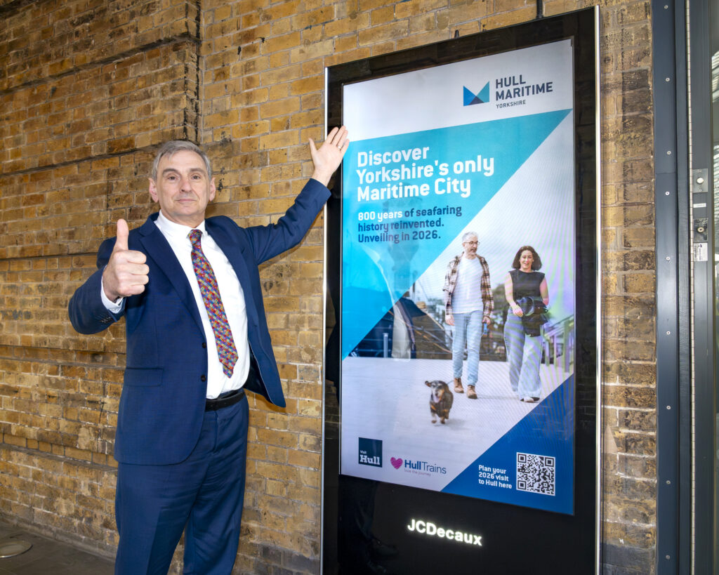 A man in a suit holds his thumb up in front of an electronic board showcasing 'Hull Maritime' artwork