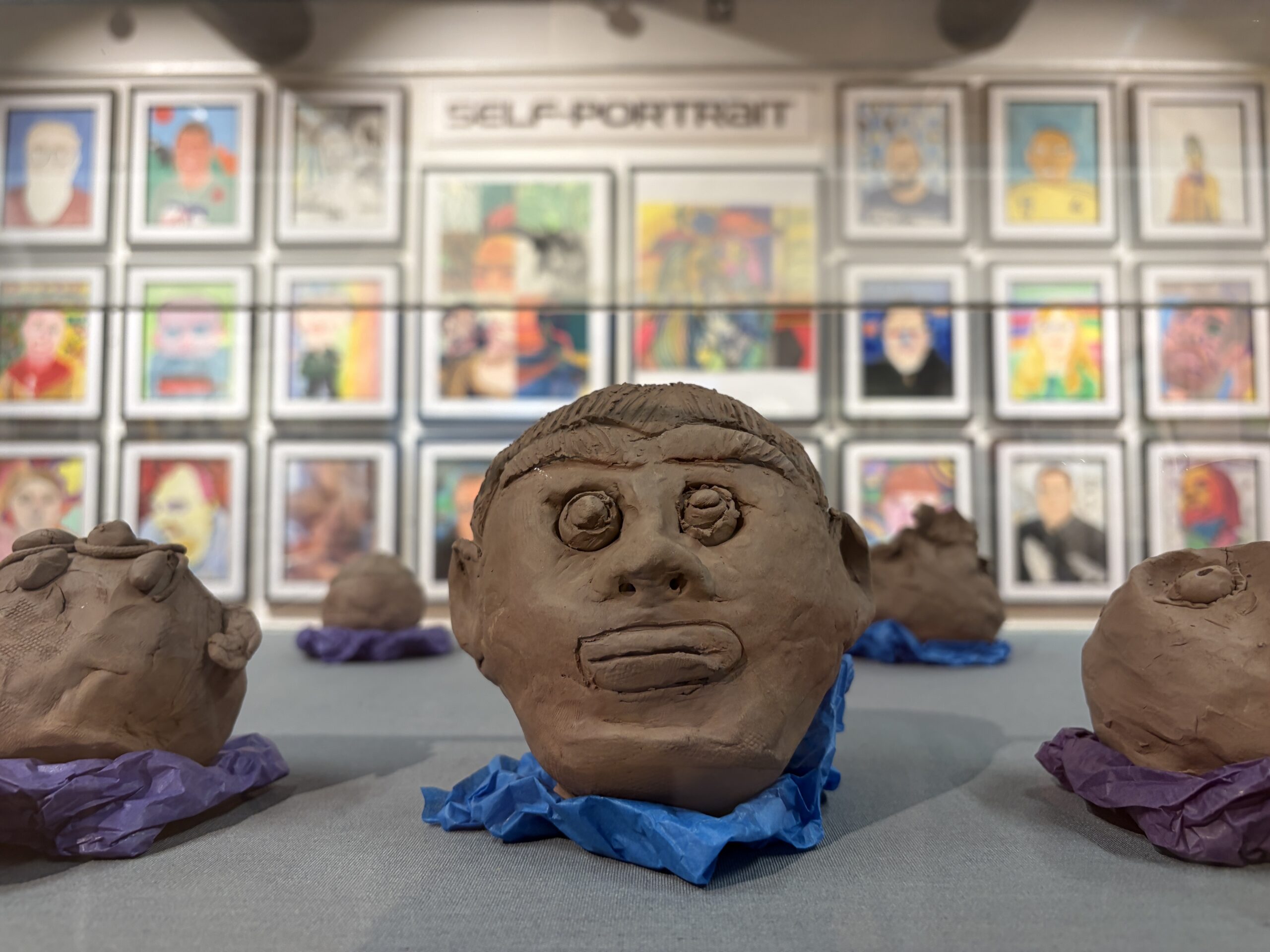 In the forefront a clay moulded head and face within a glass cabinet, with colourful portraits hung on the wall behind