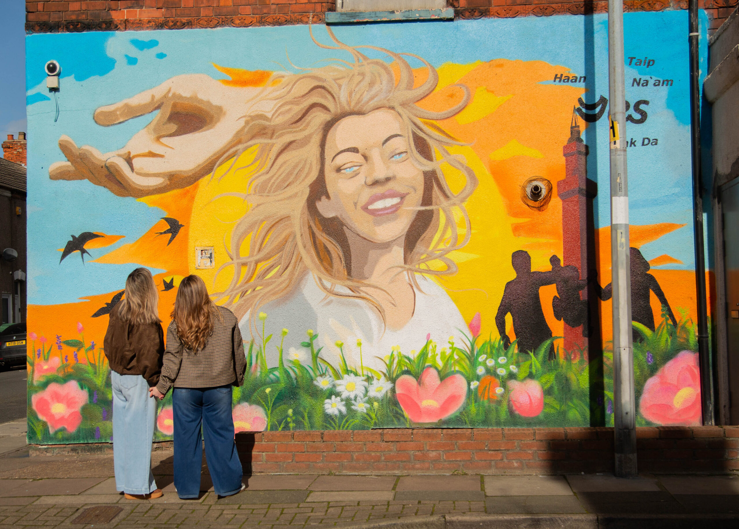 two women stand in front of a large painted mural of a smiling woman