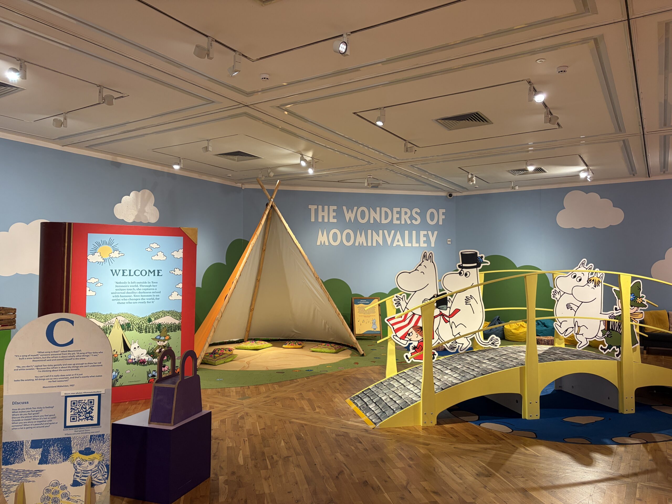 A wide shot of a room filled with a colourful exhibition including a yellow bridge and tipi tent with cushions