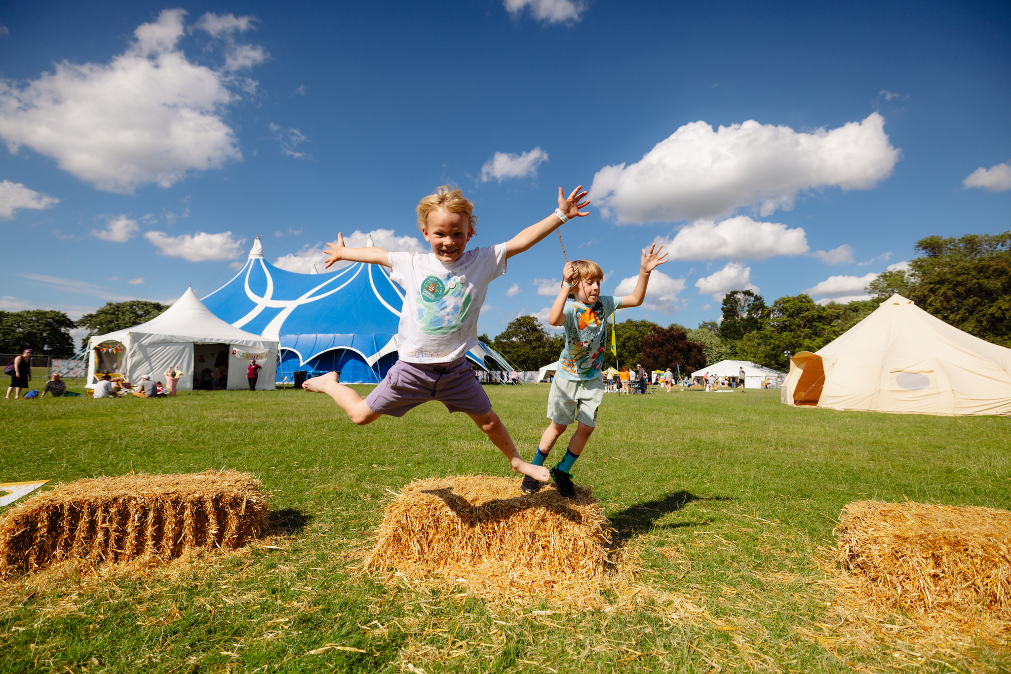 2 young children jump from a hay bale in the middle of a huge green space with circus tents in the background