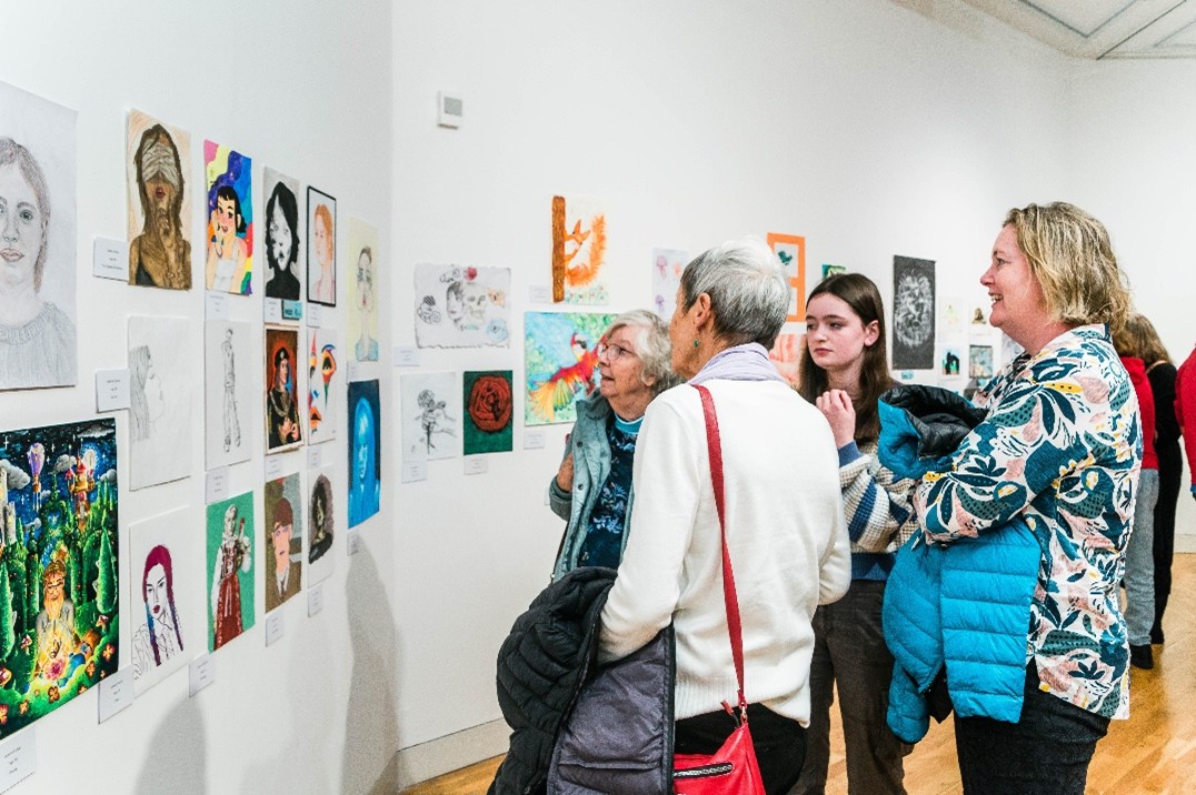 A group of people stand together in a gallery looking at paintings and pictured on a wall