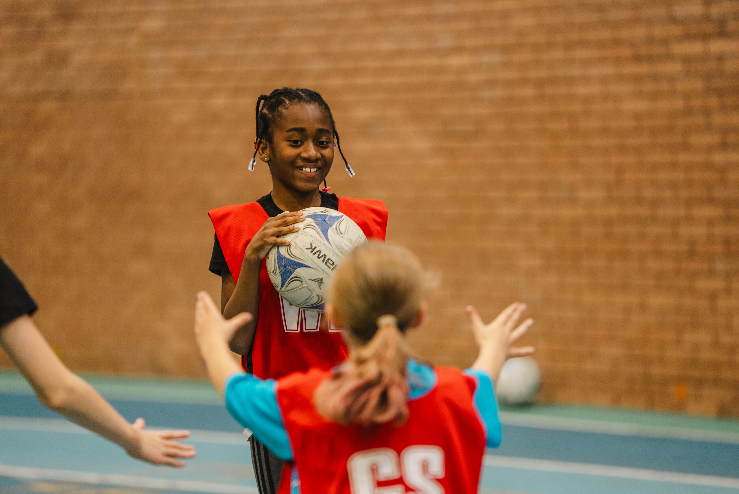 two children are playing netball. One is passing the ball to the other. They are in a sports hall.