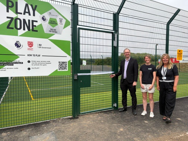 A man and two women stand in front of a gated green sports pitch with a 'Play Zone sign