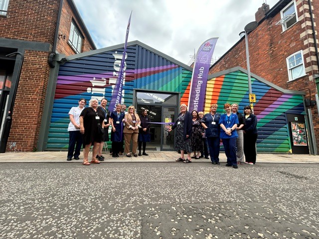 A group of people stand outside the pop-up gallery. They are preparing to cut a purple ribon. the buildinh in the background is painted in rainbow colours.
