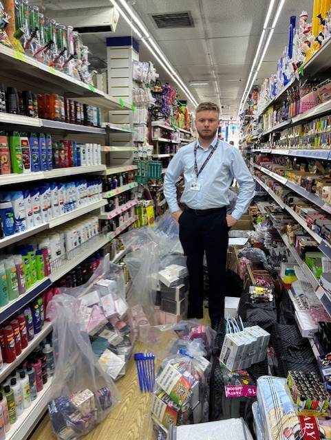 A man stands in a brightly lit aisle of a shop. He is surrounded by clear bags, filled with products being seized