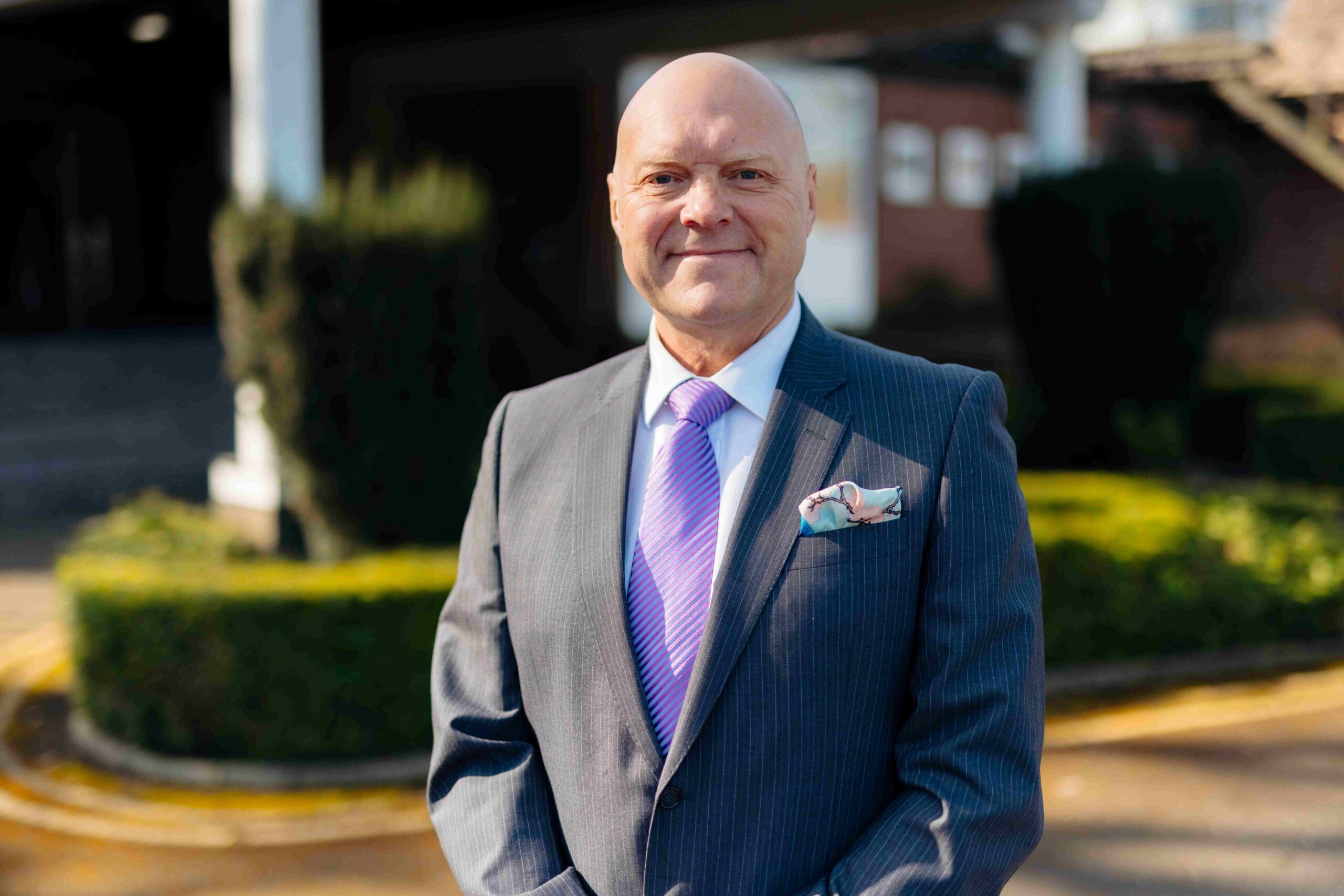 A man in a suit stands outside in the sun with greenery behind him