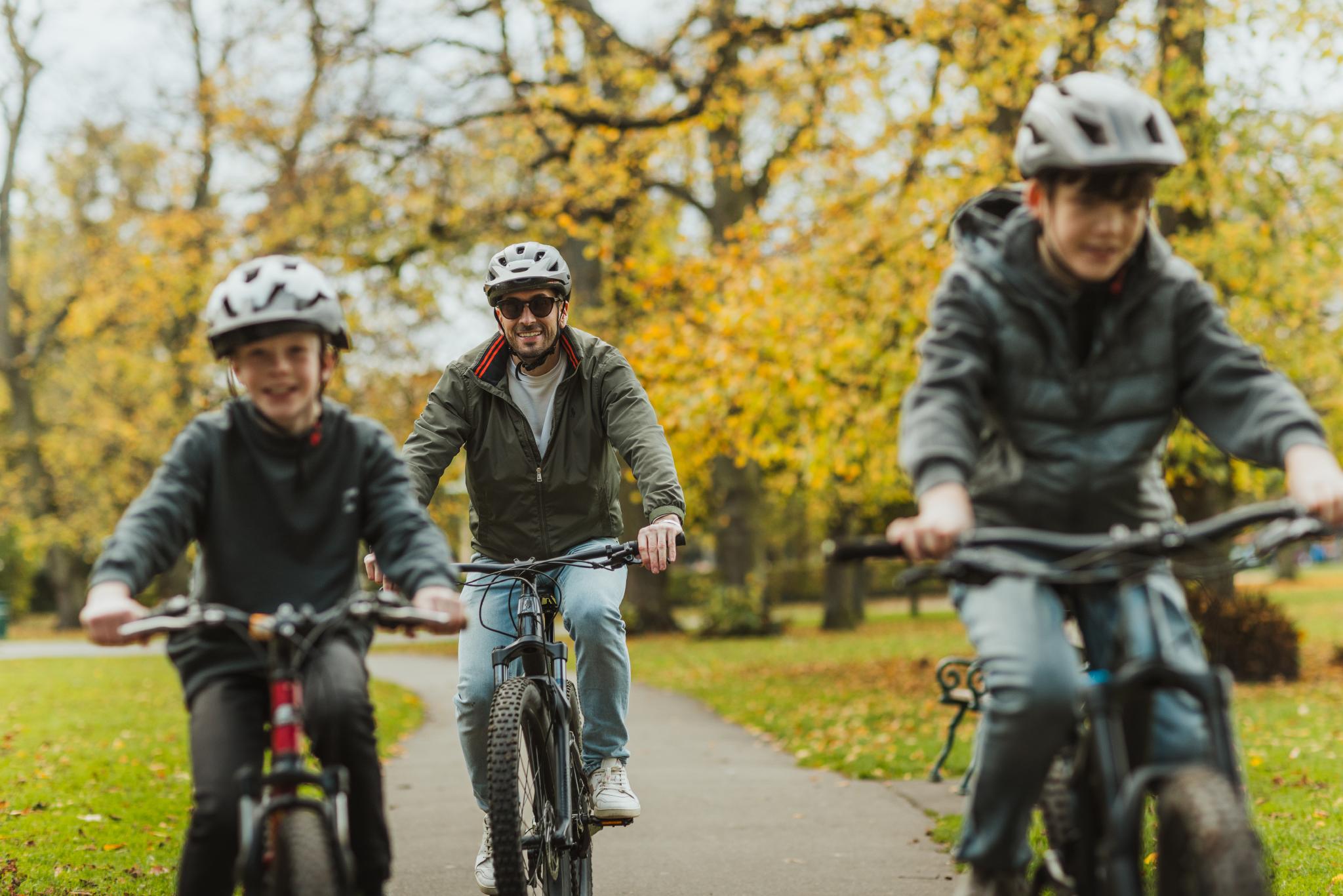 a man and two boys cycle through a park wearing helmets
