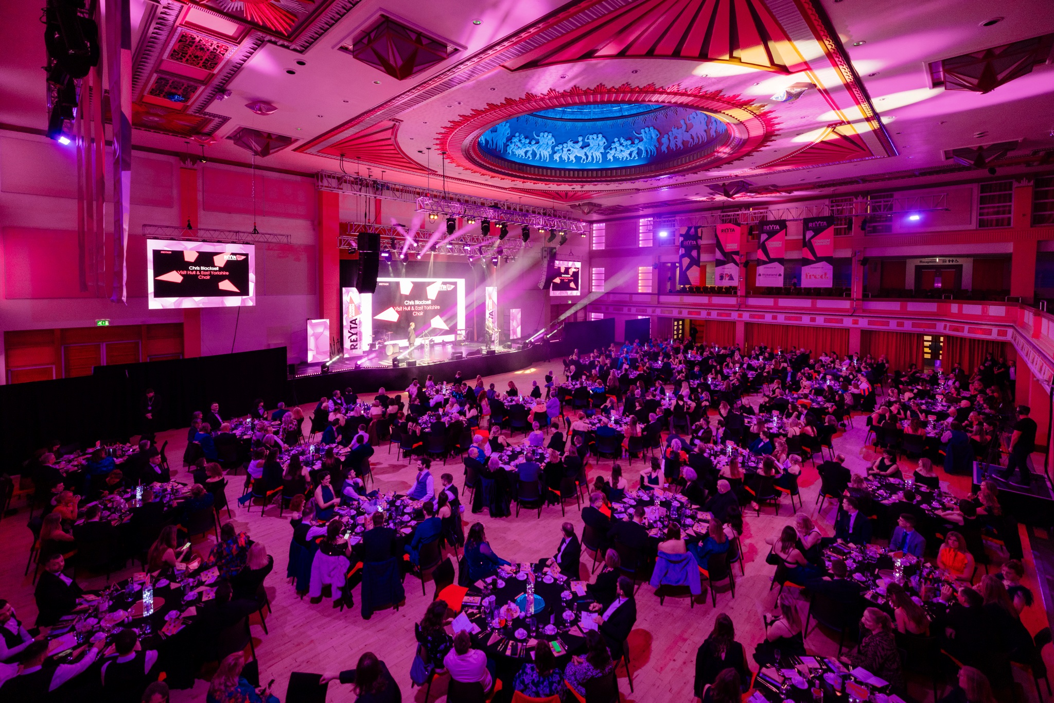 A large theatre hall is filled with round tables and people, looking at a stage with awards being presented. the room is lit pink.
