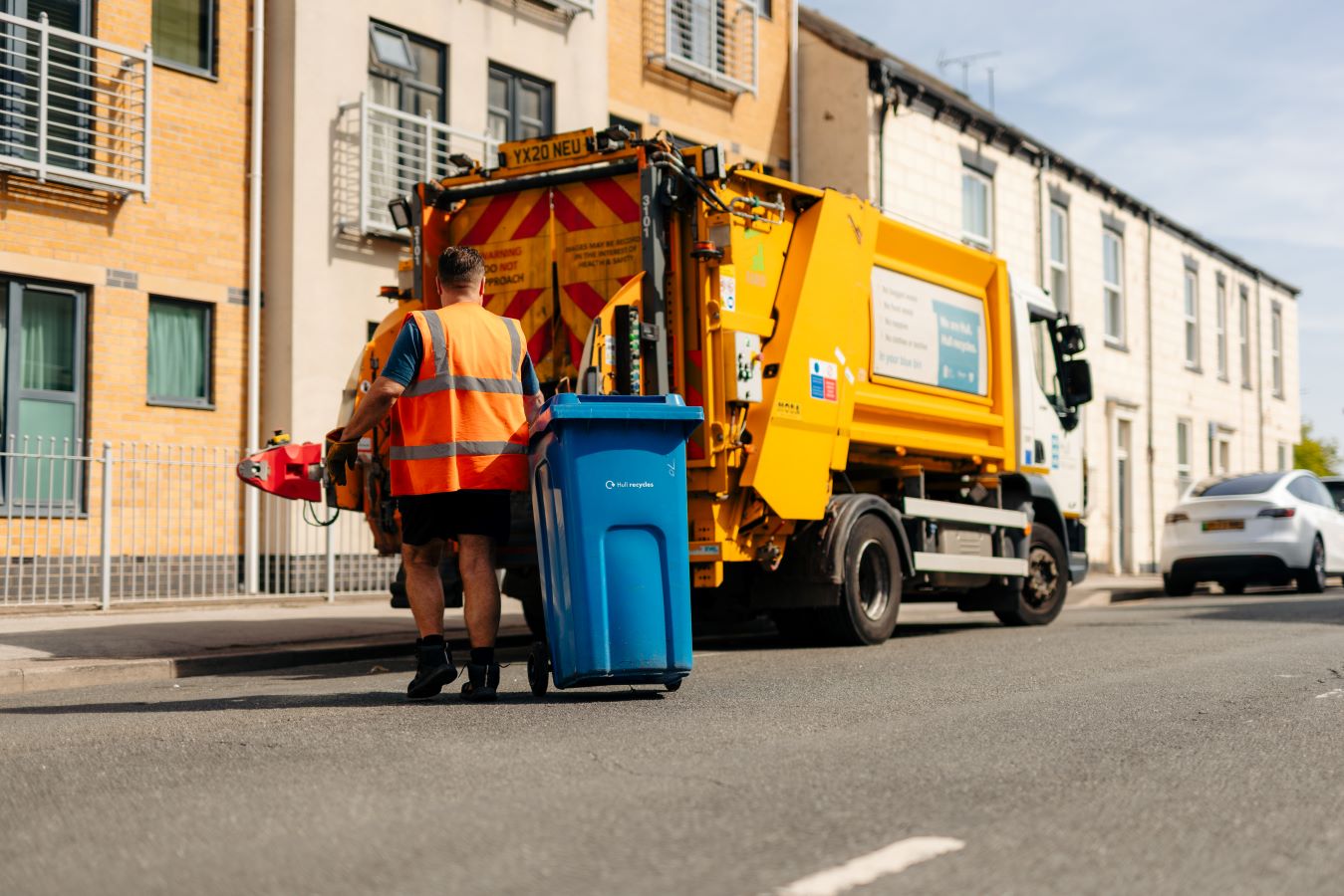 A waste operative in high-visibility clothing walking a blue recycling bin towards a waste collection vehicle for emptying.