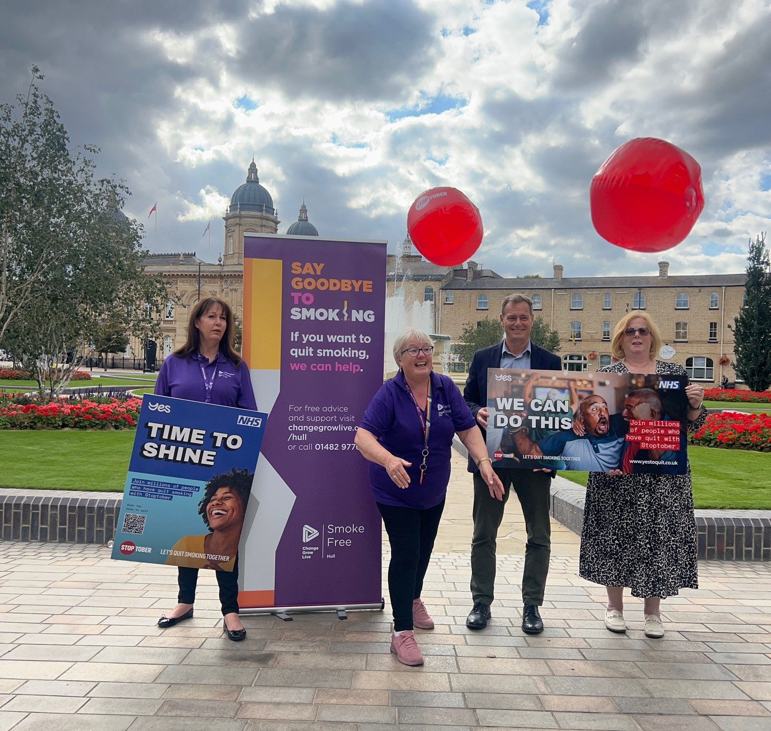 four people stand in front of flower beds, with signage promoting Stoptober. One person throws two red beach balls toward the camera (the balls represent the round, red Stoptober logo)